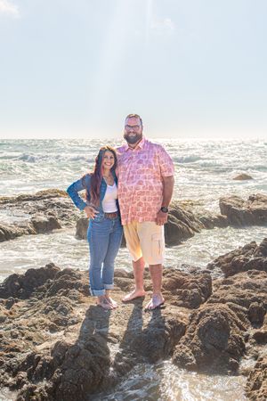 A smiling couple poses together on rugged coastal rocks as sunlit ocean waves crash behind them.