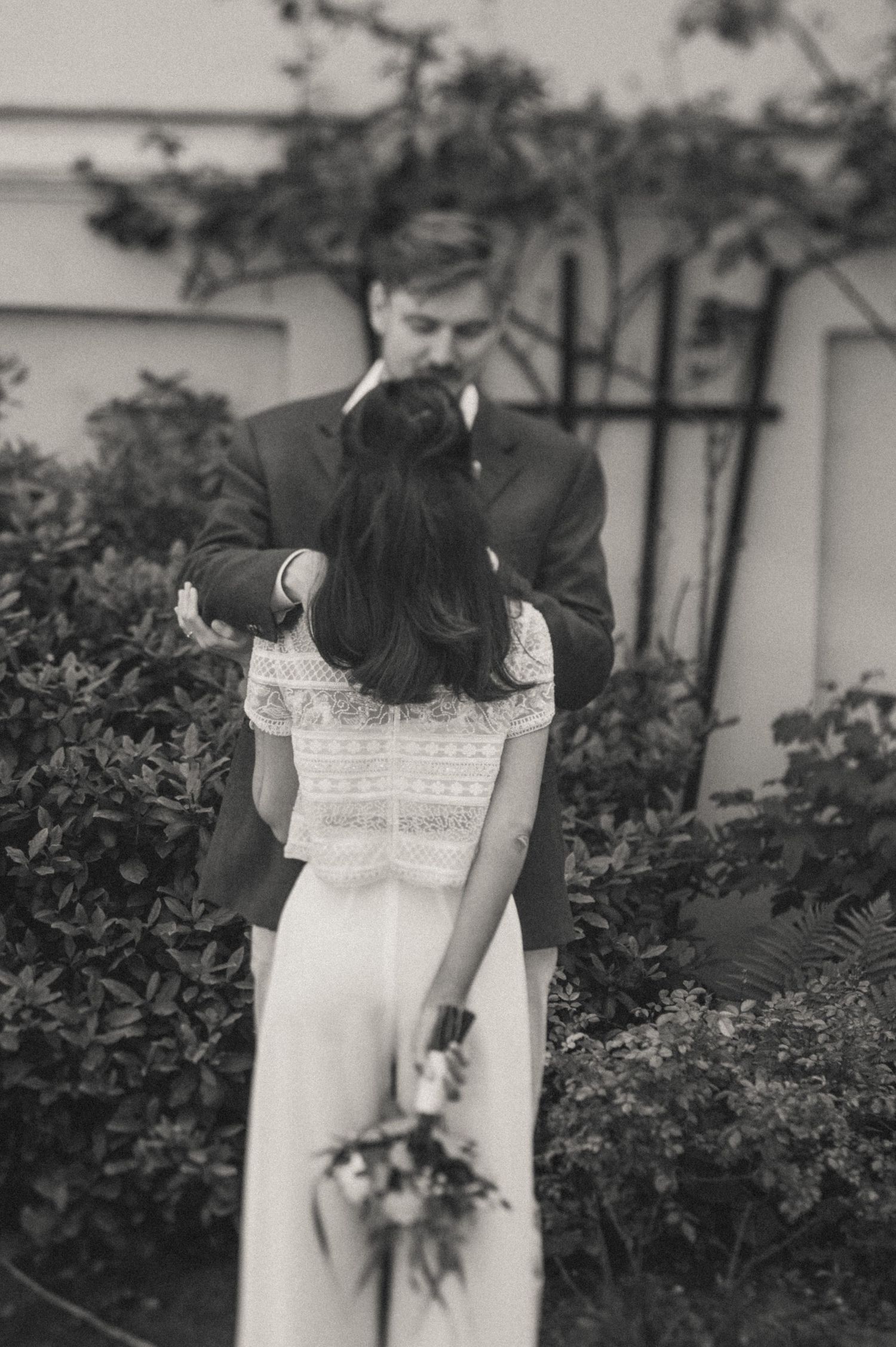 Black and white photo of a bride in a lace top holding a bouquet behind her back as she embraces a groom in a suit outdoors. The Hague