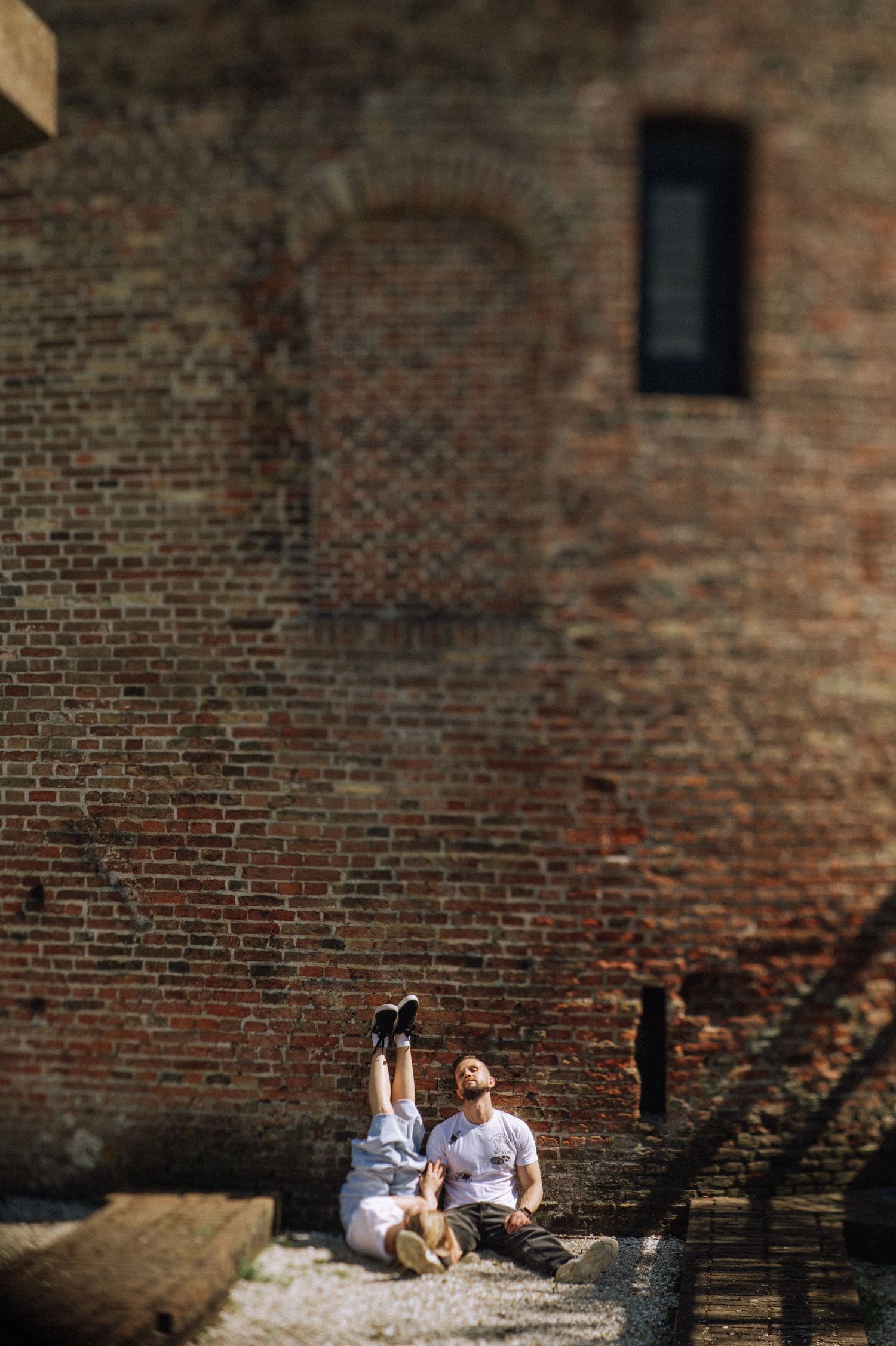Two people sit on gravel leaning against a large weathered red brick wall, Lisse, Huys Dever