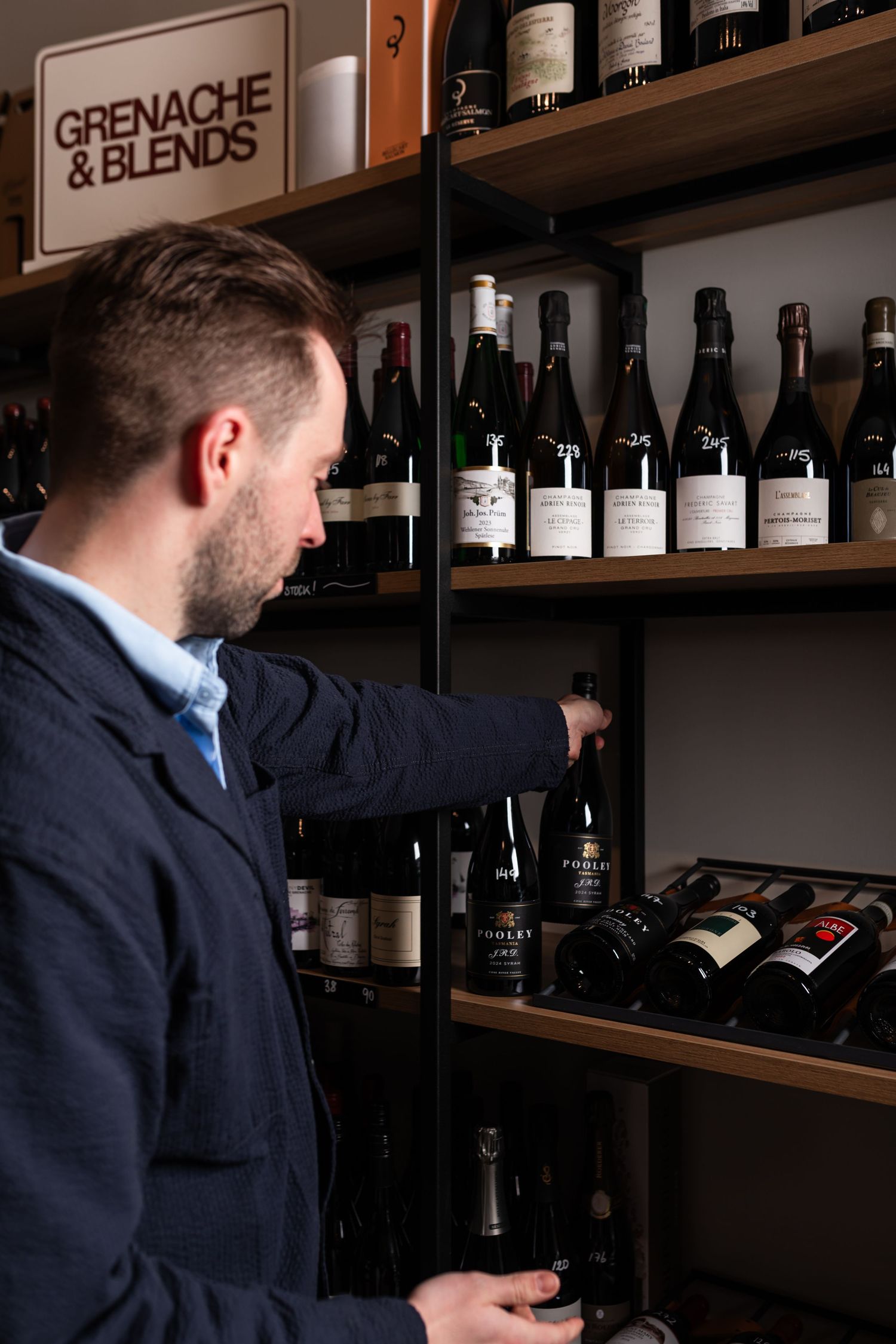 Man in a navy jacket browses bottles on a wine shop shelf beneath a Grenache & Blends sign.
