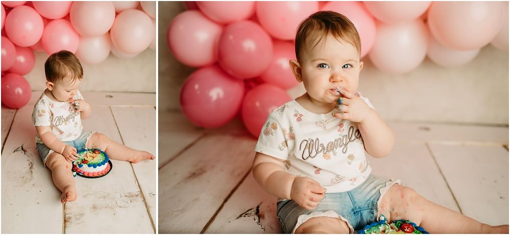 Baby enjoys first birthday cake smash session with pink balloon backdrop on white wood floor.