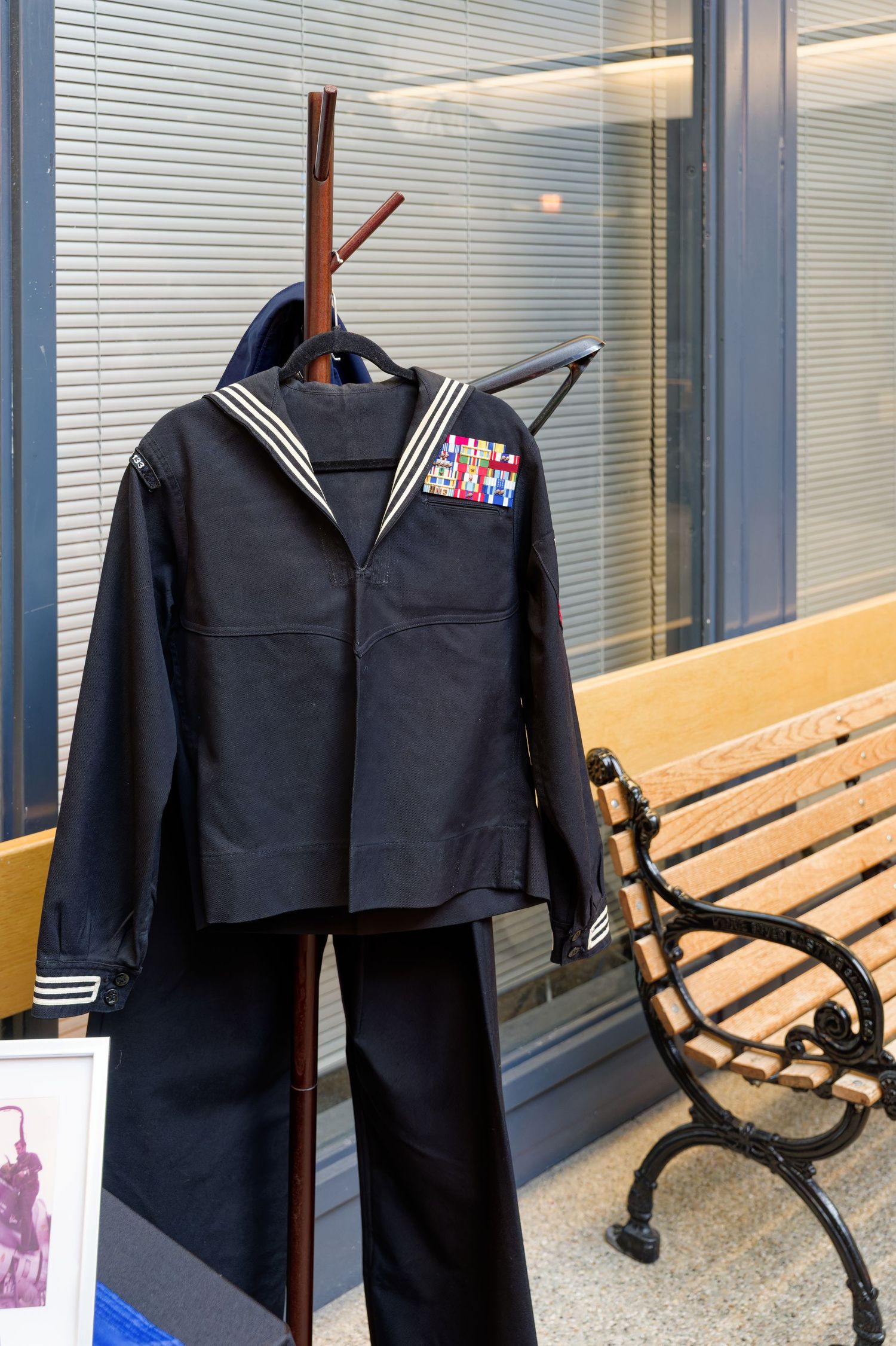 Navy enlisted dress blue uniform with medals displayed on a wooden coat rack near a bench.
