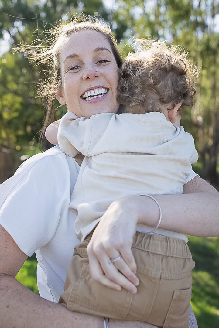 Toddler in denim overalls sits barefoot on a white blanket in the grass beside a two-layer vanilla cake with cream frosting.