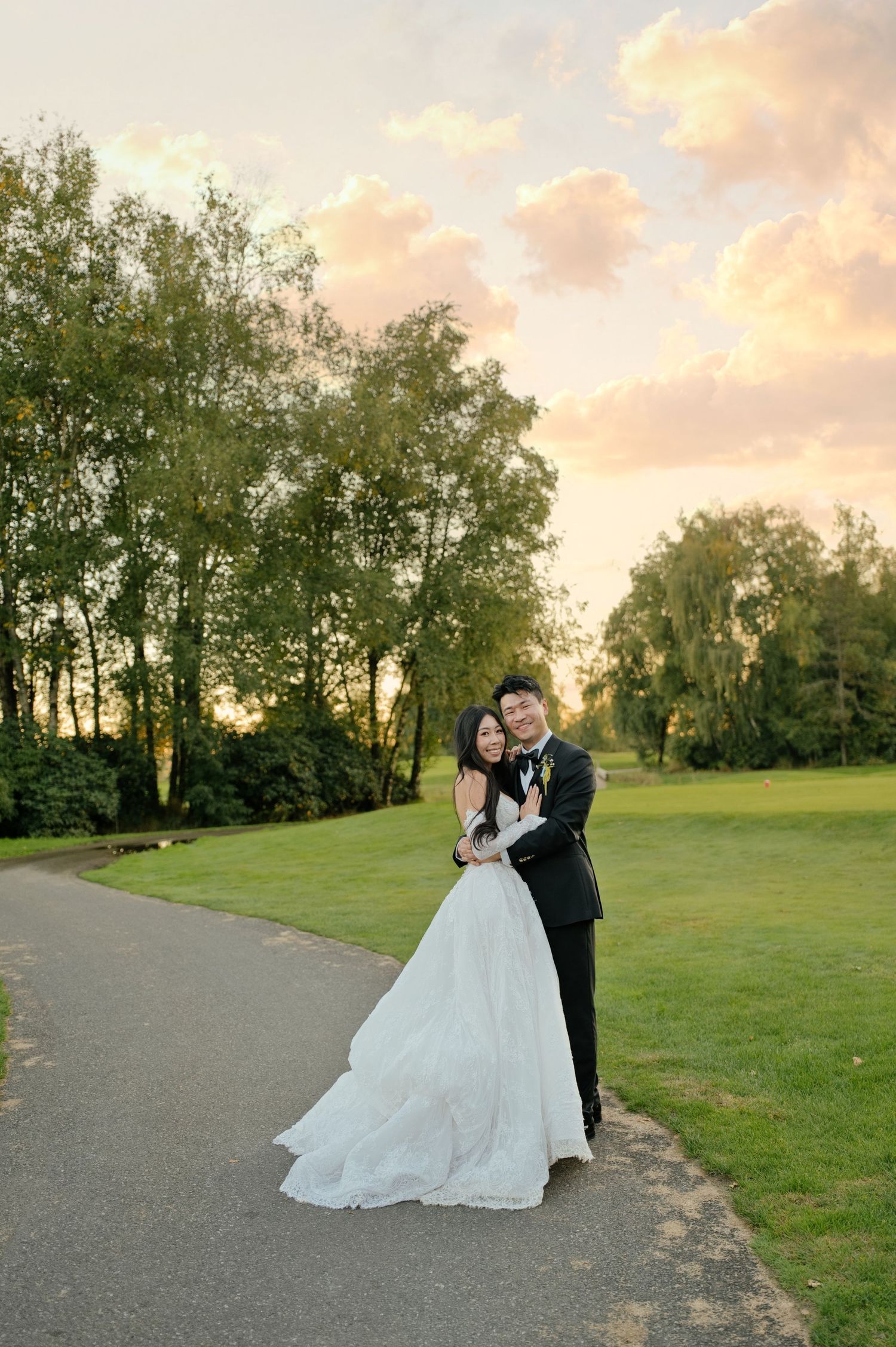 Bride in a white gown and groom in a black tuxedo embrace on a paved path beside a green golf course at sunset.