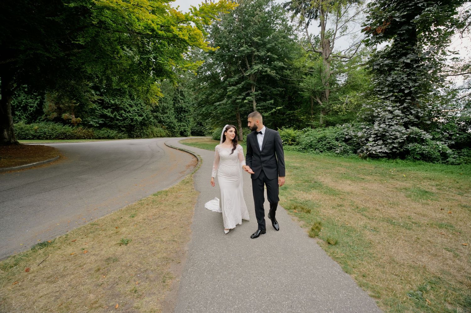 Bride in a white gown and groom in a black suit walk hand in hand along a curved park path surrounded by lush green trees.