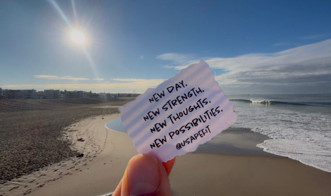 Hand holding a note reading New day, new strength, new thoughts, new possibilities against a sunny sandy beach and ocean waves.