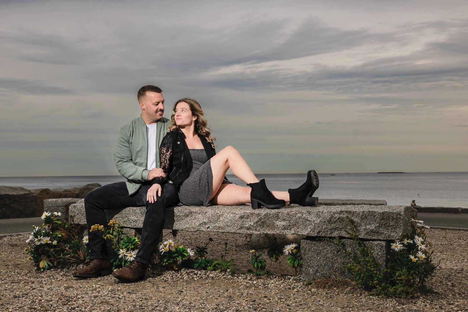 Couple sitting close together on a stone bench, gazing at each other with affection by the water.  Engagement at Rockport Harbor, Rockport,