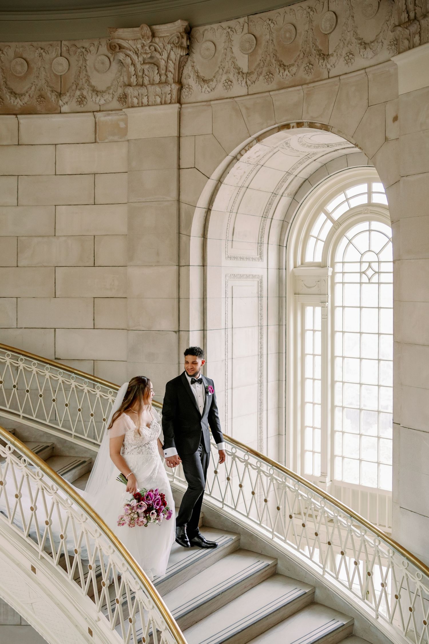 Bride and groom walking down elegant marble staircase in grand wedding venue with arched windows.