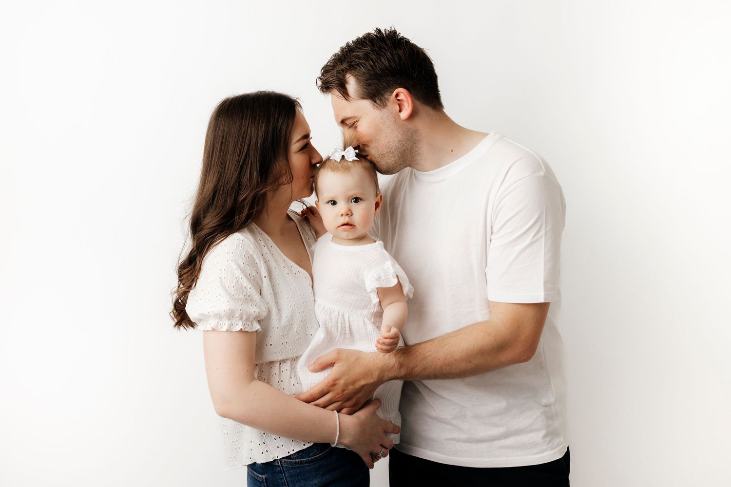 A young family in white tops poses against a white background, parents kissing their baby girl on the cheeks.