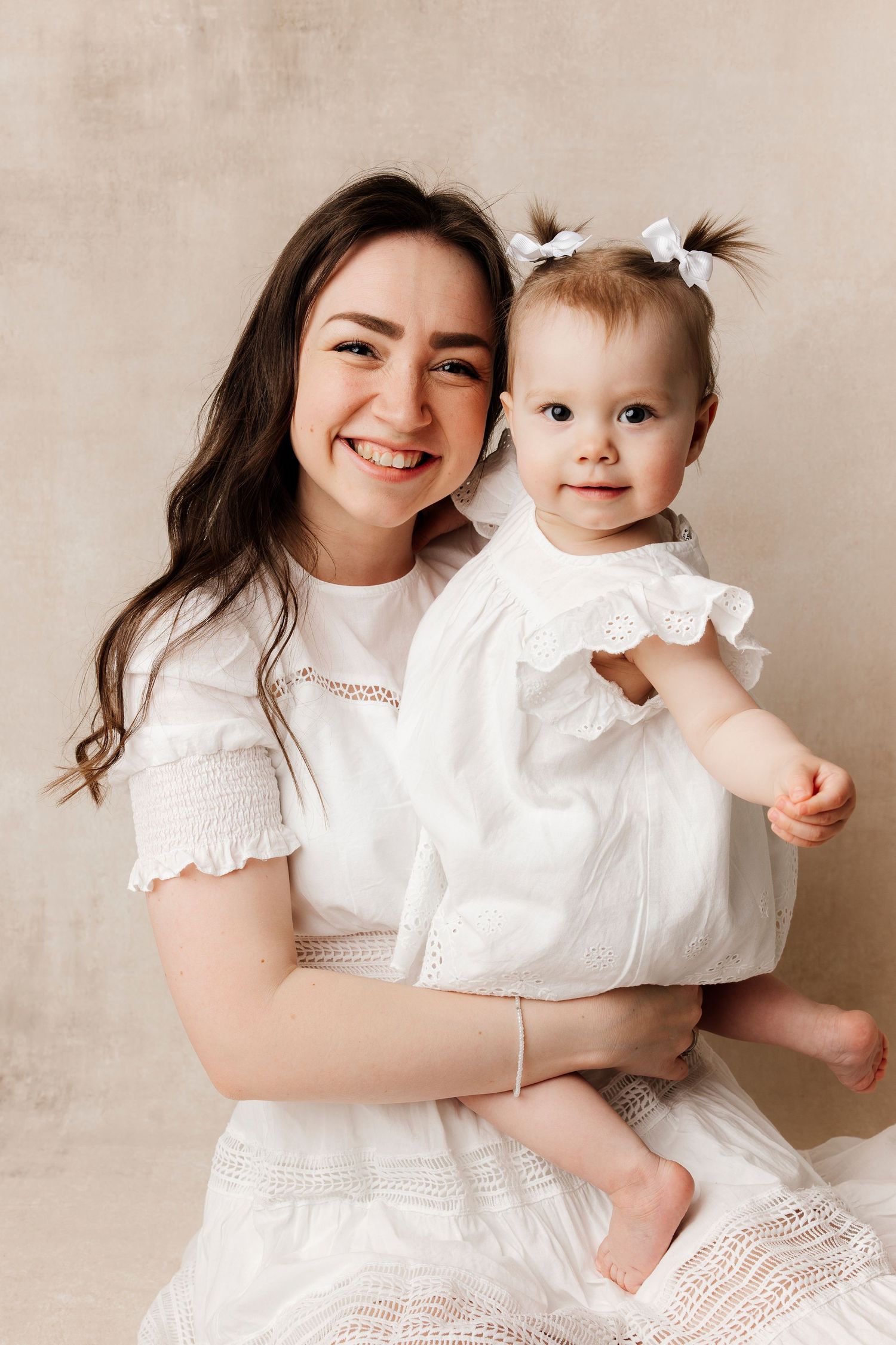 A smiling young woman in a white dress holds a cute baby girl wearing a white dress with bow pigtails in a studio.
