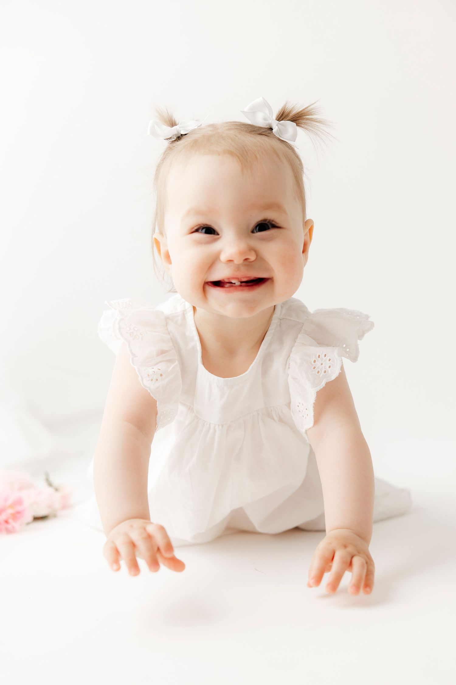 A cheerful baby girl in a white ruffled dress with pigtail bows smiles brightly while crawling on a white background.