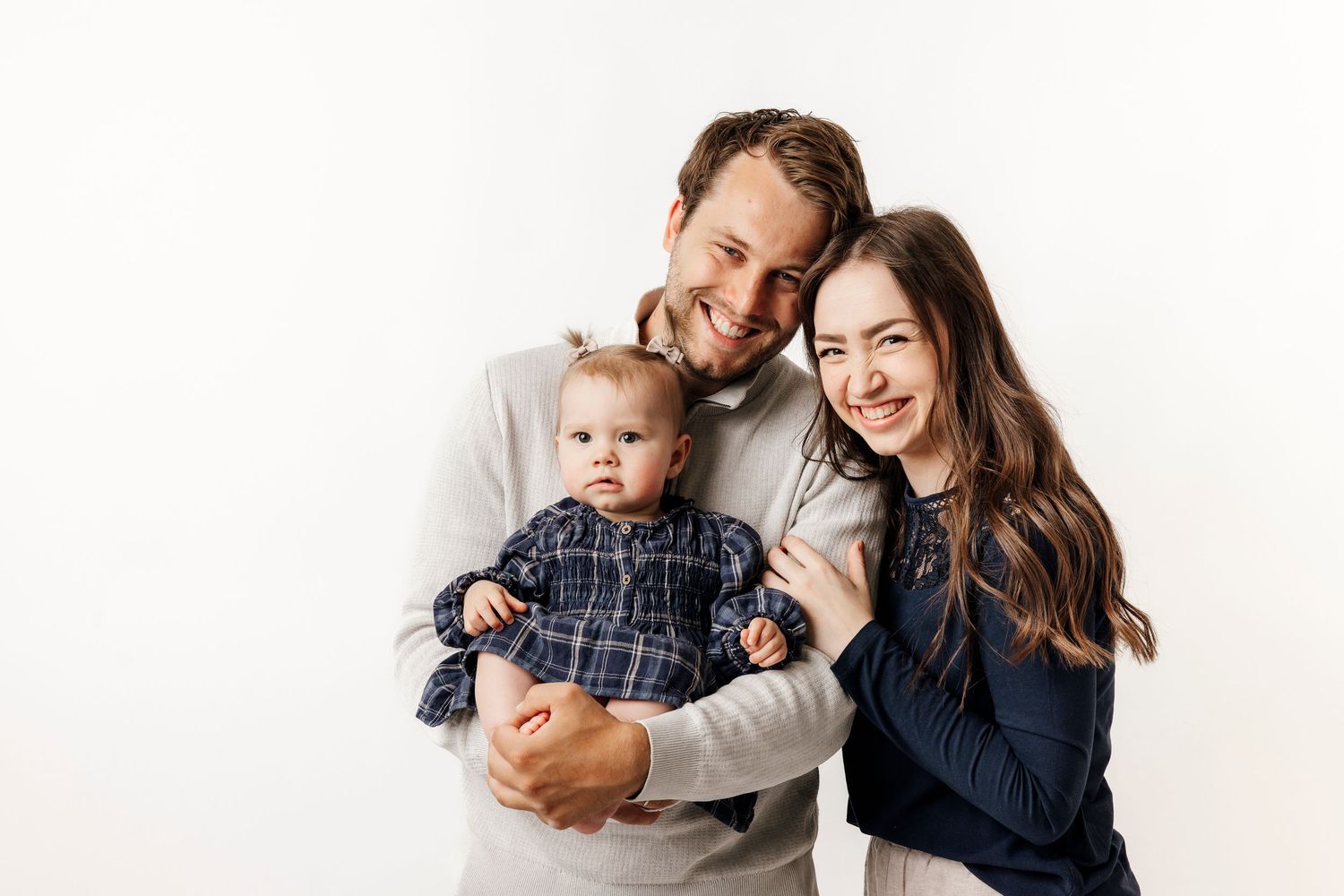 Happy young family with parents smiling and holding their baby dressed in plaid against white background.