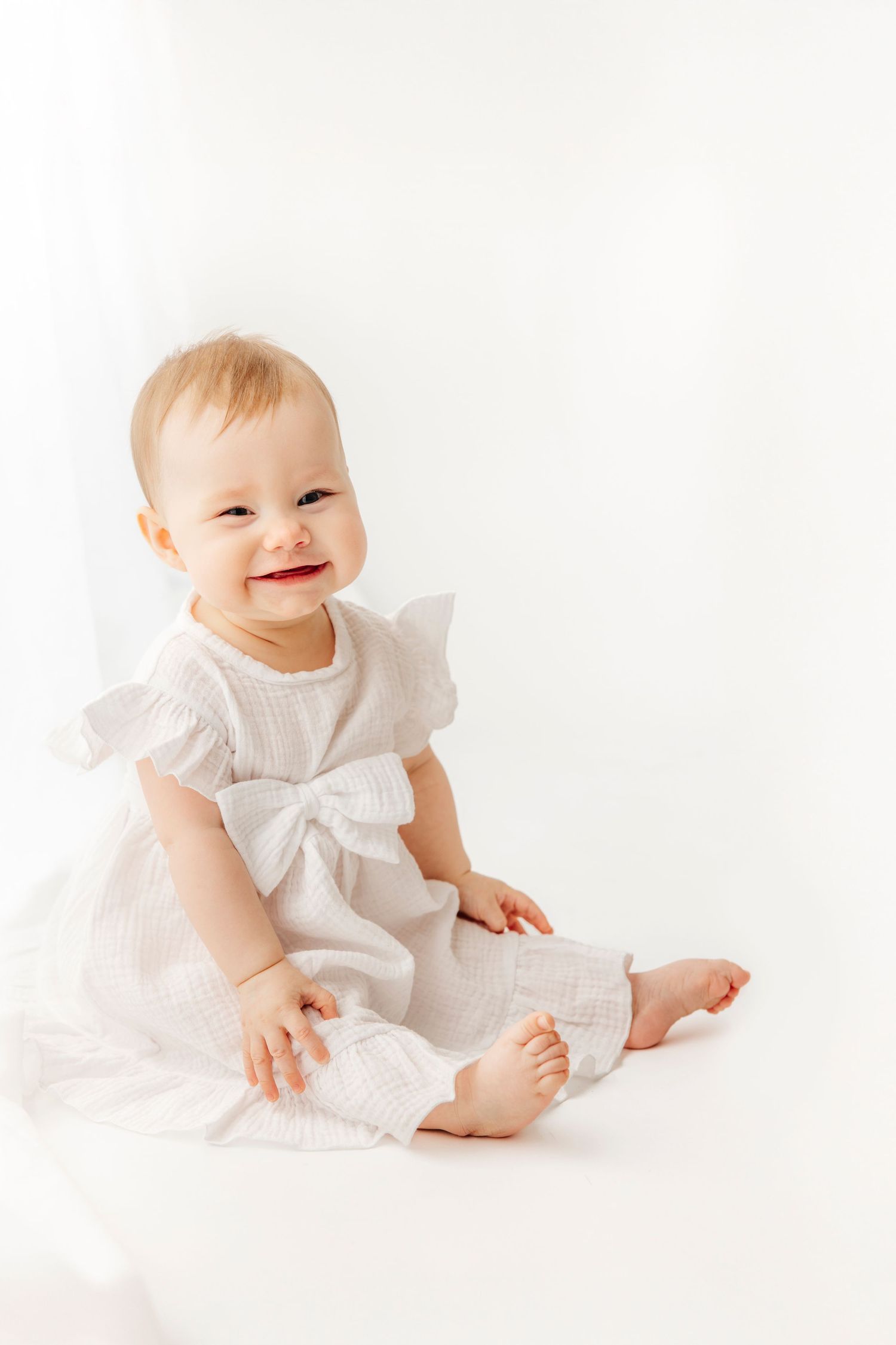 Smiling baby girl in white dress with bow sits barefoot on white background.