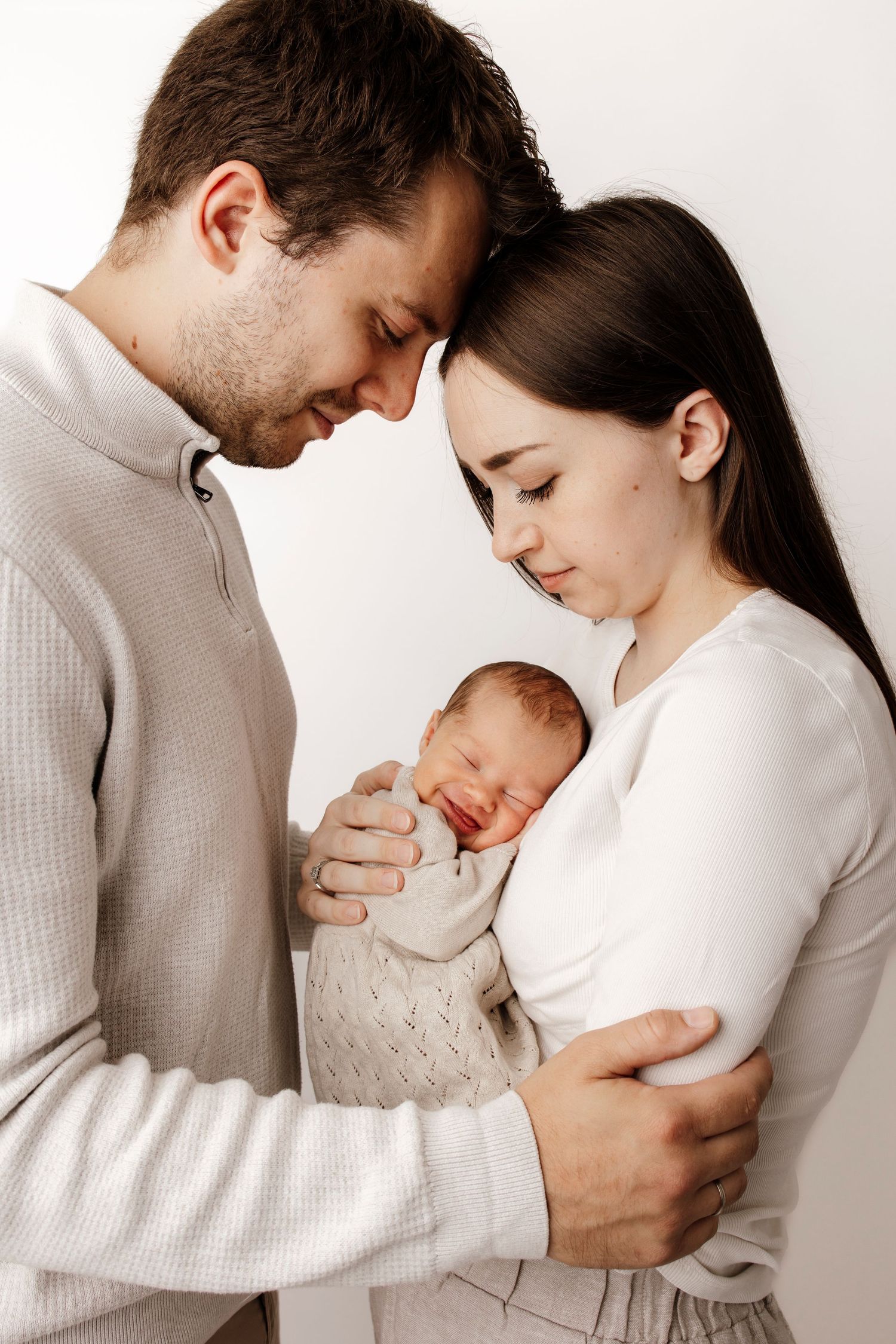 New parents tenderly holding their sleeping newborn baby, with foreheads touching in an intimate family portrait.