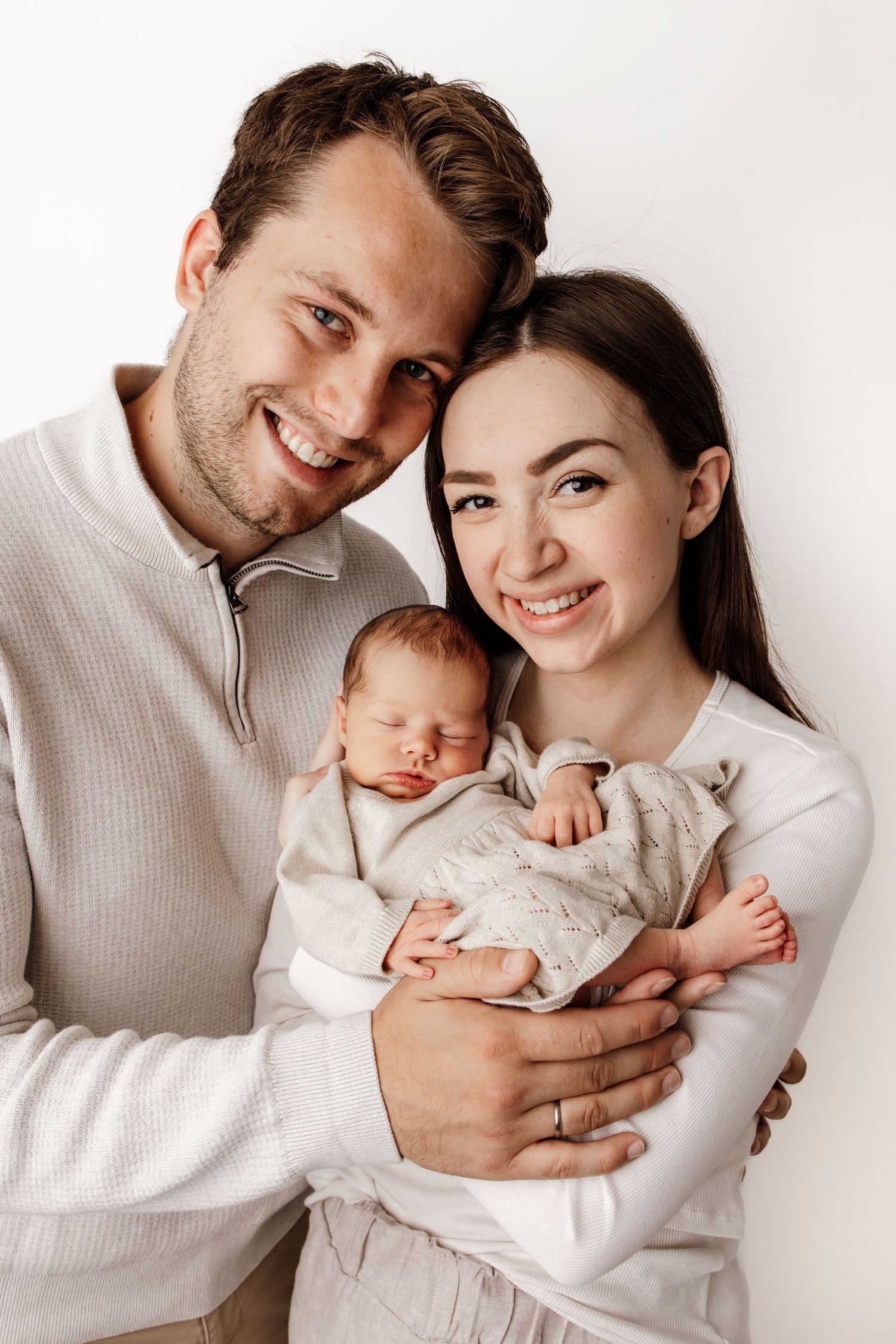 Happy young parents smiling and holding their sleeping newborn baby, dressed in neutral tones against a white background.