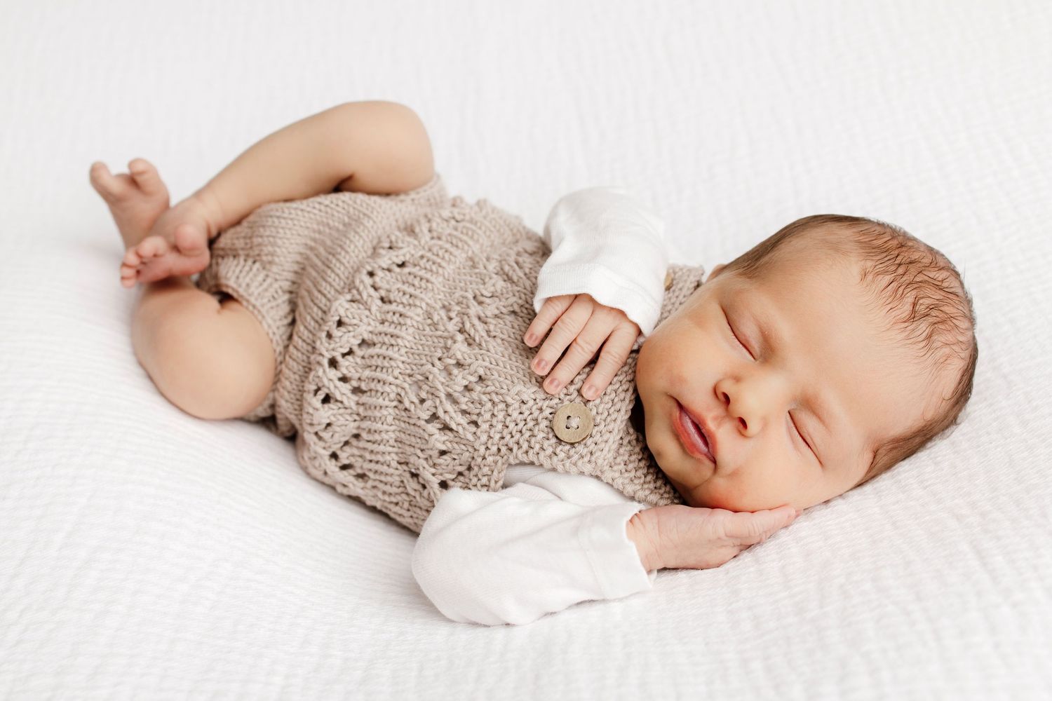 Peaceful newborn baby sleeping in a knit beige romper with white mittens on a soft white blanket.