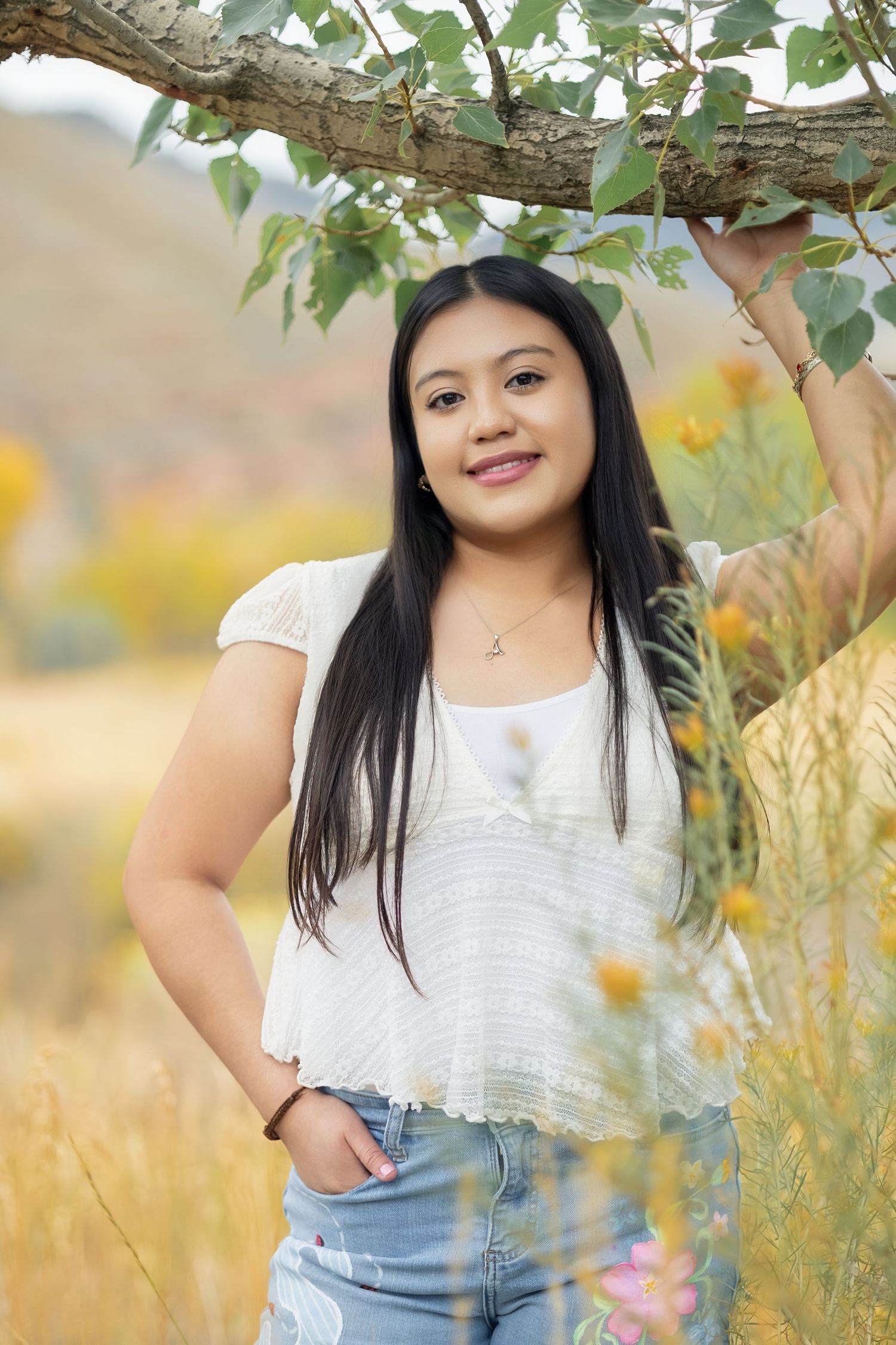 Young woman in white lace top and jeans smiles outdoors near a tree branch with autumn foliage.