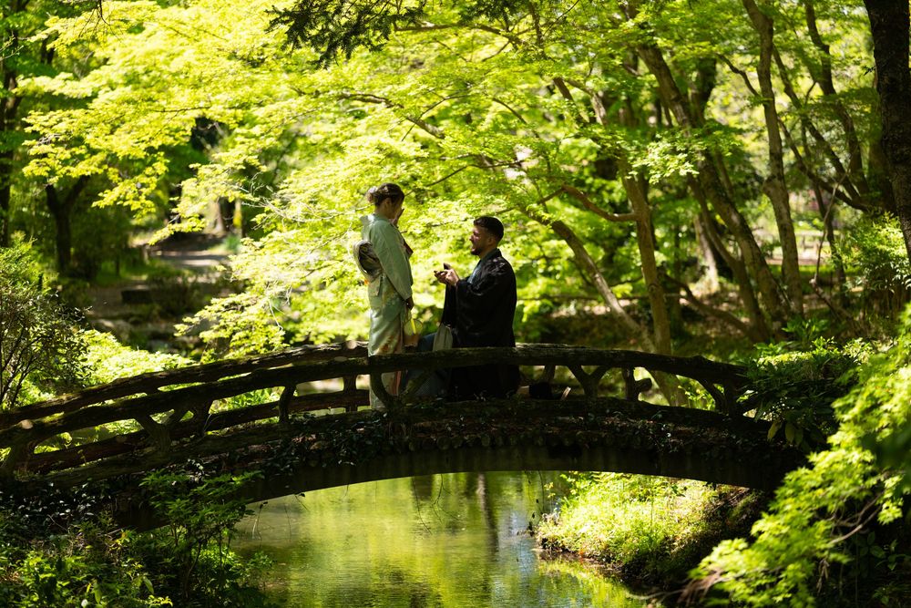 Couple in traditional Japanese attire stands on an arched wooden bridge surrounded by lush green maple trees above a calm garden pond.