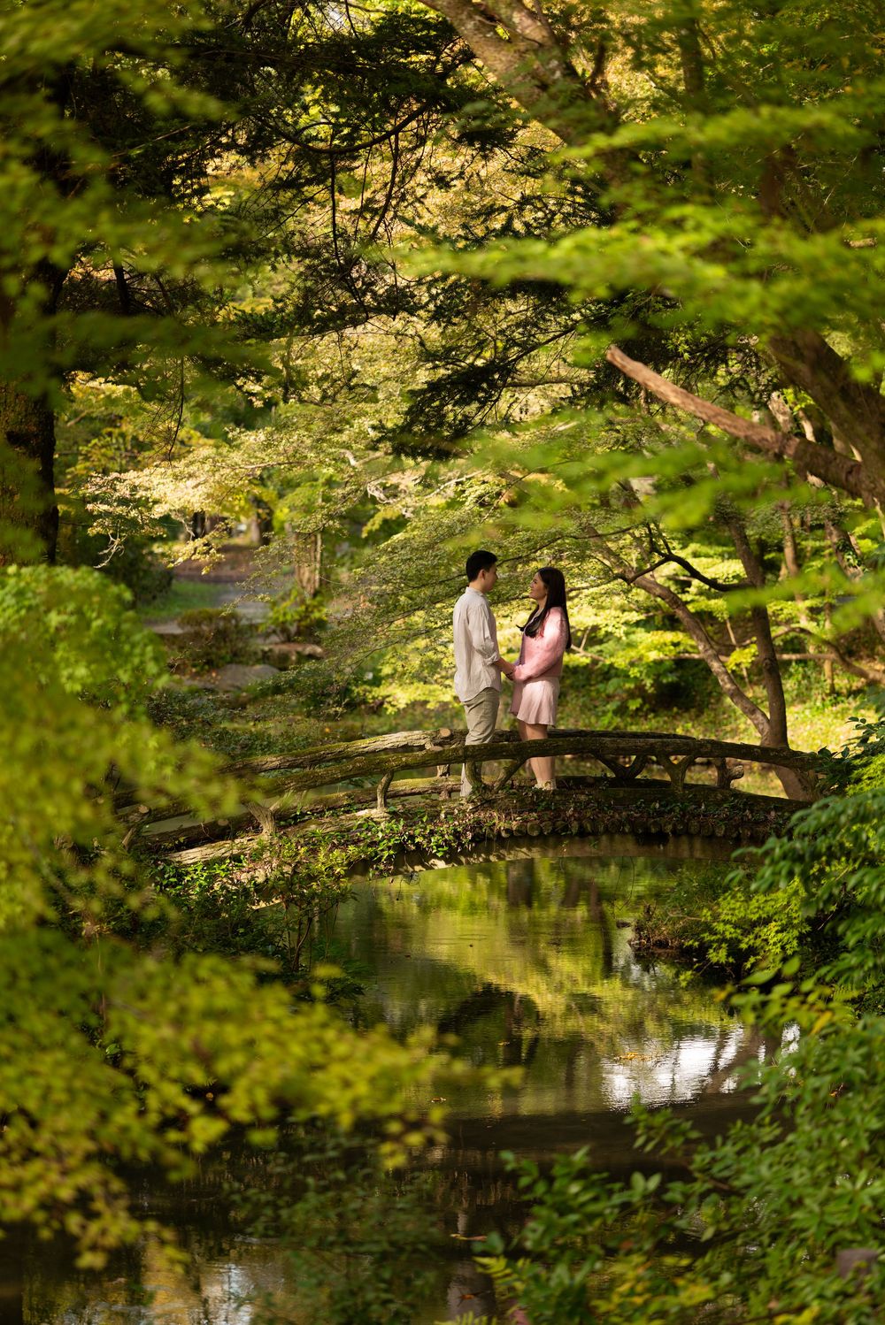 Couple standing face to face on an arched wooden bridge over a calm pond, surrounded by lush green trees in a serene Japanese-style garden.