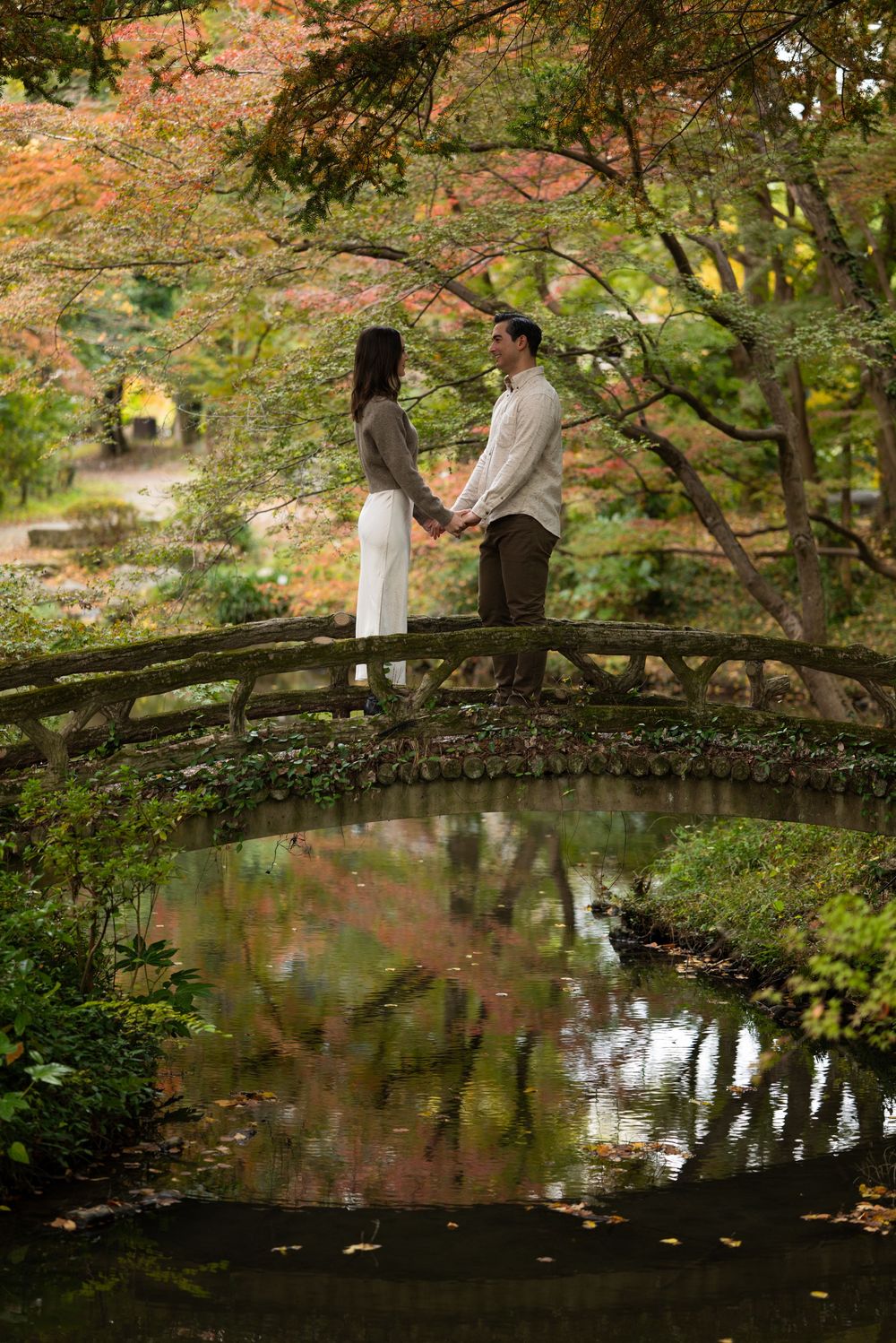 Couple holding hands on a rustic wooden bridge in a Japanese garden, surrounded by colorful autumn foliage reflected in the pond below.