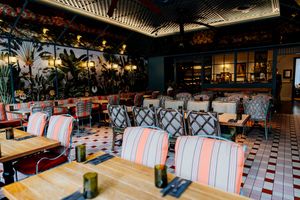 Stylish restaurant interior with pink striped chairs, wooden tables, checkered tile floor, lush tropical plants and warm ambient lighting.