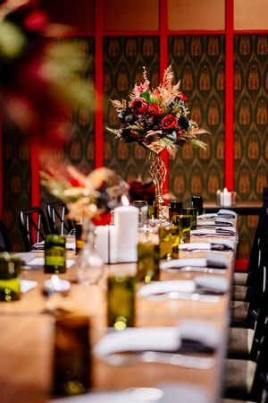 Long wooden banquet table set with white napkins, green glass votives, candles and a tall autumn floral centerpiece in a patterned red-accen