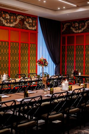 Elegant banquet room with long wooden tables, black chairs, candlelight, red floral centerpieces and ornate red and green patterned walls.