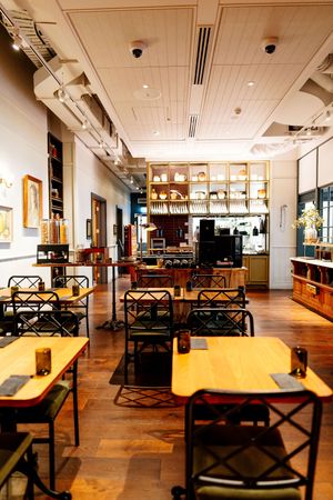 Bright, modern restaurant interior with wooden tables, green cushioned chairs, hardwood floors, and a open kitchen with shelves of dishware.