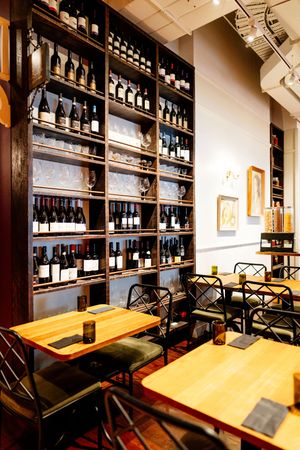 Cozy wine bar interior with wooden tables, black metal chairs, and tall shelves displaying rows of wine bottles and glassware.