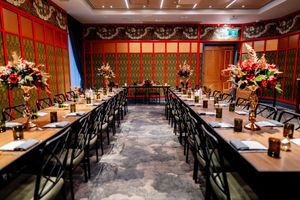 Elegant banquet hall with long wooden tables, black chairs, red floral centerpieces and ornate red walls with gold patterned trim.
