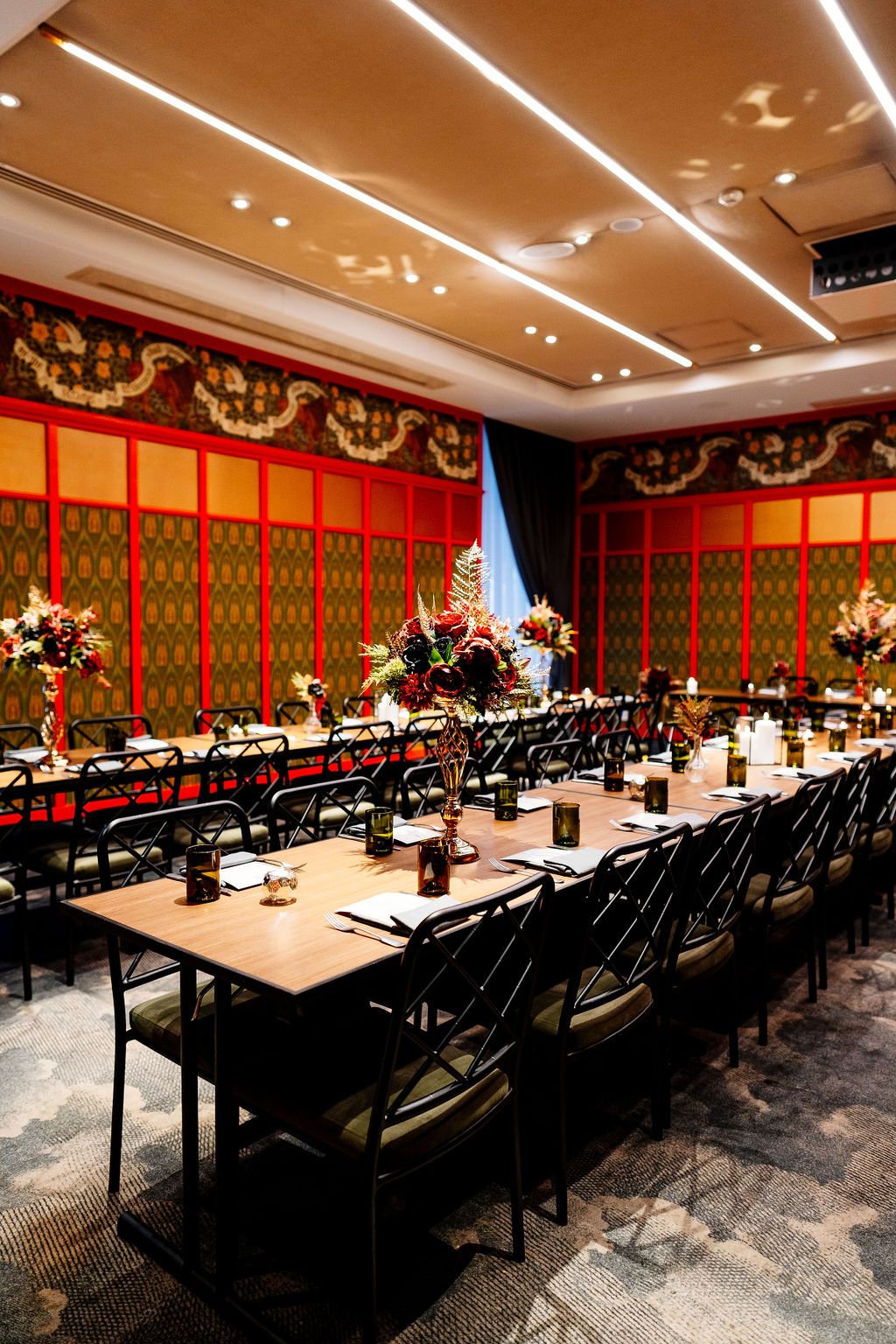 Elegant banquet room with long wooden tables, black chairs, floral centerpieces and red paneled walls with ornate gold patterns.