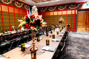 Long banquet table set with green glass goblets, menus, and a tall red floral centerpiece in an ornate dining room with red and green walls.