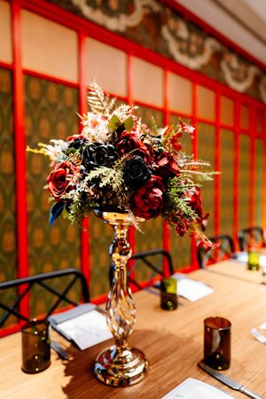 Elegant gold pedestal vase with dark red and burgundy roses and greenery as a wedding table centerpiece against ornate red-framed walls.