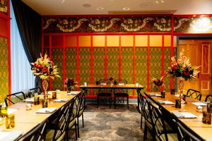Elegant private dining room with red lacquered walls, gold floral panels, long tables, black chairs and vibrant flower arrangements.