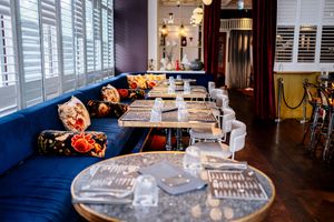 Elegant restaurant interior with a blue velvet banquette, floral throw pillows, set tables, white chairs, and shuttered windows.