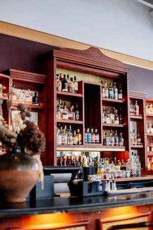Well-stocked bar with rows of liquor bottles on wooden shelves, a dark countertop, and a vase of dried flowers in the foreground.