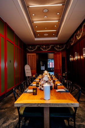 Long wooden private dining table set with candles, floral centerpieces and black chairs in an elegant room with red trim and dark wood walls