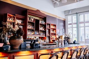 Elegant bar interior with a wooden back shelf of bottles, a lit counter, empty stools, and large windows letting in natural daylight.