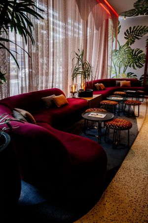 Plush burgundy velvet curved sofas, round tables and patterned stools in a stylish lounge with tropical leaf wallpaper and sheer curtains.