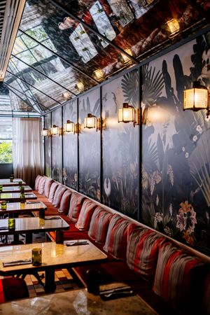 Elegant restaurant interior with striped coral banquettes, wooden tables, glowing brass lanterns, tropical botanical wall mural and mirrored