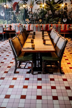 Long wooden restaurant table with green cushioned rattan chairs on a red and white tiled floor, beneath a tropical botanical mural.