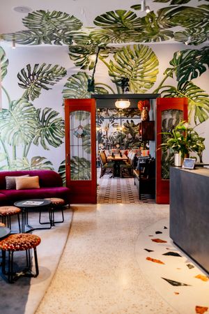 Stylish lounge with large tropical leaf murals, a burgundy velvet sofa, terrazzo floors and red-framed glass doors leading to a dining room.
