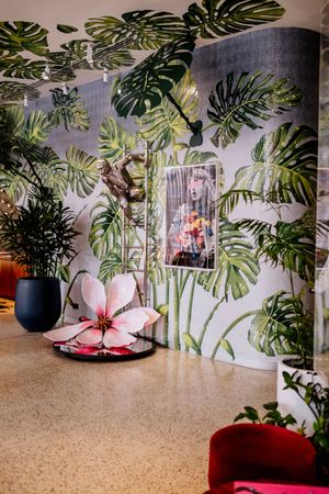 Lobby with tropical monstera leaf wallpaper, a large pink magnolia sculpture, a framed portrait, potted plants and a red velvet chair.