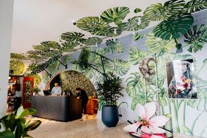 Bright hotel lobby with a hand-painted tropical leaf mural, potted plants, a pink orchid and a reception desk where staff stand.