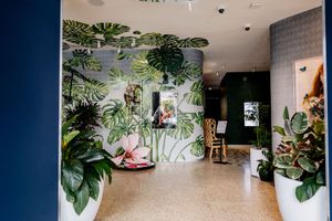 Modern lobby with a hand-painted monstera leaf mural on a curved wall, potted tropical plants, terrazzo flooring, and a wooden chair.