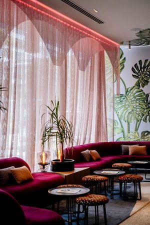 Stylish lounge with curved burgundy velvet banquettes, round side tables, spotted stools, sheer pink curtains and a tropical leaf mural.