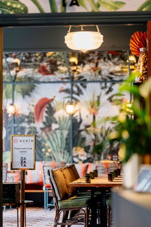 Zata restaurant interior with wooden tables, green chairs, a tropical botanical mural, a pendant lamp and a please wait to be seated sign.