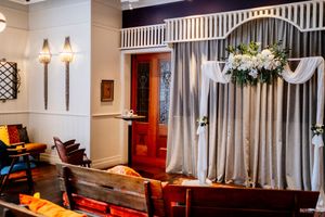Elegant indoor wedding ceremony space with draped gray curtains, white floral arch, wooden pews, wall sconces and a red paneled door.