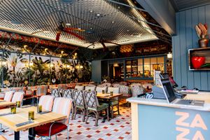 Bright restaurant interior with patterned tile floors, wooden tables, red and green chairs, tropical plants, and a blue ZAZA counter.