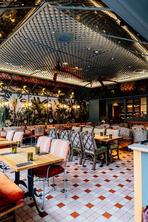 Tropical-themed restaurant interior with orange striped chairs, wooden tables, checkered tile floor and a patterned lattice ceiling.